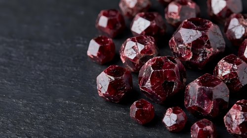 Sparkling red garnet gemstones with faceted cuts scattered on dark slate surface for jewelry making and luxury accessories design projects.