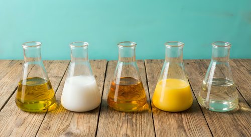Five glass laboratory flasks containing different colored liquids including milk, oils and clear solutions arranged on rustic wooden surface for scientific research.