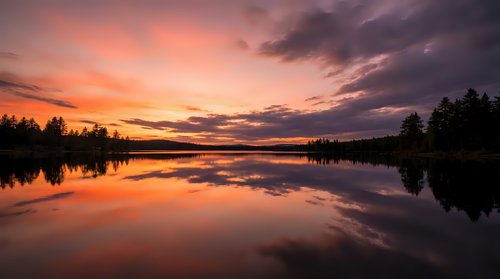 Serene lake at sunset with dramatic orange and purple sky reflecting on calm water, surrounded by silhouetted evergreen trees along shoreline.