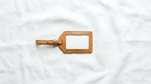 Brown leather luggage tag with blank white label on marble surface for travel identification and personalization.