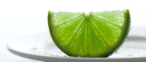 Fresh lime slice on white plate with water droplets, citrus fruit segment for beverage, cooking, and food styling projects.