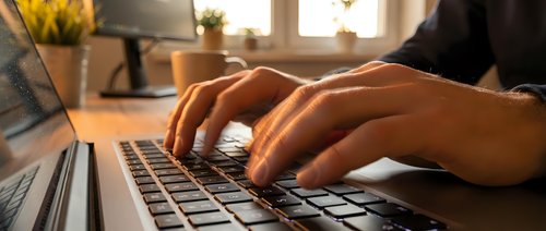 Close-up of male hands typing on a laptop keyboard at a wooden desk, warm sunlight in background, remote work concept.