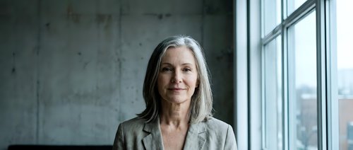 Mature woman with long gray hair and fair complexion wearing casual beige cardigan, standing by large window with urban cityscape view, natural daylight portrait for business, corporate, or lifestyle use.