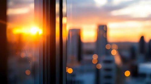 Blurred urban cityscape at sunset with bokeh lights through window glass, creating atmospheric mood with golden hour glow and reflections. Modern architecture silhouettes.