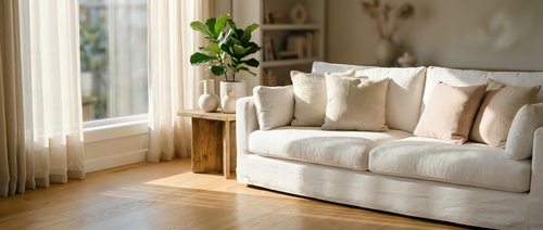 Modern bright living room interior with white sofa, wooden floors, and natural light streaming through windows creating cozy home atmosphere.
