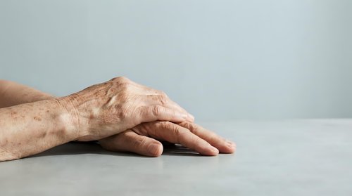 Elderly hands with visible veins and wrinkles resting on white surface against neutral background showing aging process and senior healthcare concepts.