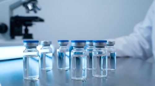 Glass vials with blue caps arranged on a lab table, microscope and scientist in white coat blurred in background.