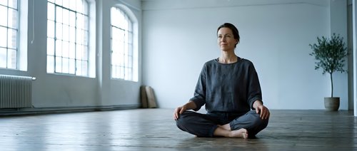 Young Caucasian man practicing meditation in lotus position on wooden floor in bright modern loft studio with large windows and minimalist interior design.