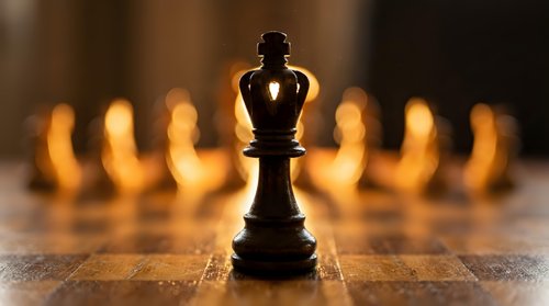 Black chess king piece standing prominently on wooden board with blurred golden chess pieces in background creating dramatic lighting and strategic game atmosphere.