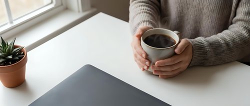 Woman in cozy sweater holding coffee mug at white desk with laptop and plant, perfect for remote work and home office lifestyle concepts.