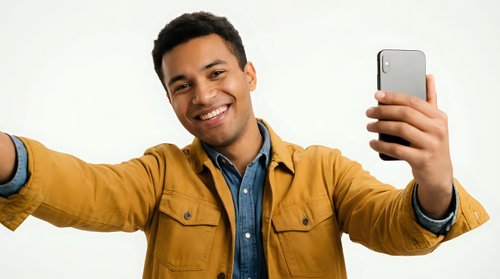 Young man with dark hair and medium brown skin tone wearing mustard yellow jacket over denim shirt taking selfie with smartphone, arms outstretched, smiling at camera against white background for social media and technology concepts.