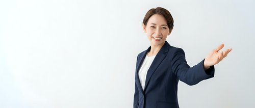 Professional Asian businesswoman in navy suit presenting with welcoming gesture against white background for corporate marketing materials.