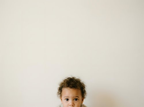 Young child with curly brown hair and light brown skin looking at camera against neutral beige wall background, suitable for parenting, childcare, education, and family-related content.