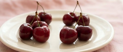 Fresh ripe red cherries with stems arranged on white ceramic plate against soft pink textile background for healthy eating and summer fruit concepts.