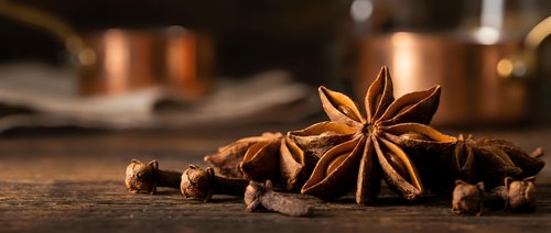 Star anise and cloves scattered on rustic wooden surface with warm copper cookware in blurred background creating cozy kitchen atmosphere for culinary designs.