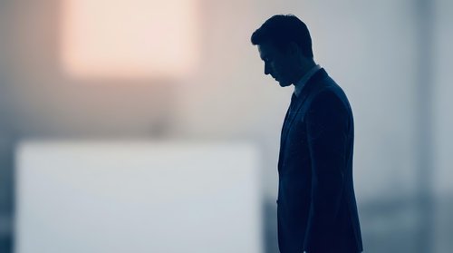 Young adult male in dark business attire standing in contemplative pose against blurred urban background. Suitable for corporate, mental health, depression awareness, and professional lifestyle concepts.