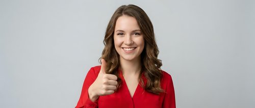 Young professional woman in red blazer giving thumbs up gesture with confident smile against neutral background for business success concepts.