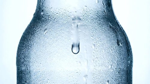 Closeup of cold water droplets condensing on frosted glass bottle surface with single drip falling, isolated on white background for beverage, refreshment and hydration design projects.