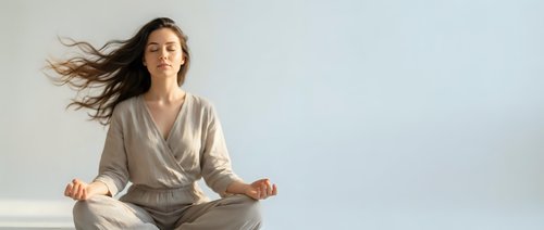 Young woman with flowing hair meditating in lotus position wearing beige outfit against white background. Peaceful mindfulness practice for wellness and relaxation.