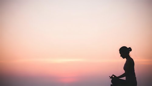 Young woman meditating in lotus pose during sunset with peaceful pink sky background for wellness and mindfulness practices.