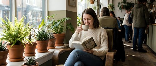 Young Asian woman reading book while sitting by window with potted plants in cozy cafe. Relaxed student enjoying literature in modern coffee shop interior.