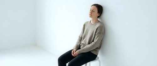 Young Asian woman in beige sweater sitting thoughtfully on white chair against minimalist background for lifestyle and wellness content.