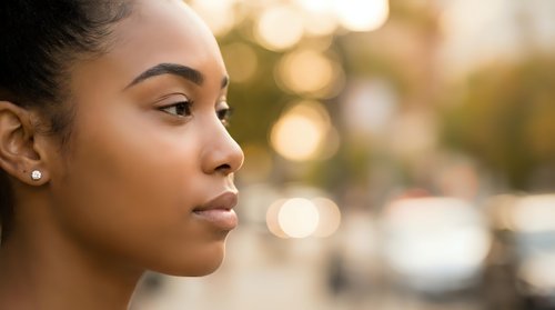 Young African American woman in profile with natural makeup and earrings outdoors with warm bokeh lighting background for lifestyle and beauty concepts.