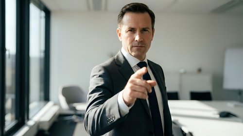 Confident middle-aged Caucasian businessman in dark suit pointing at camera in modern office environment for corporate leadership concepts.
