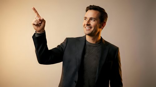 Confident young Caucasian businessman in dark suit pointing upward with finger, smiling at camera against neutral background for corporate presentations.