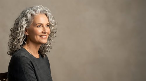 Mature woman with long gray curly hair wearing dark sweater, smiling at camera against neutral beige background. Portrait for advertising, healthcare, lifestyle.
