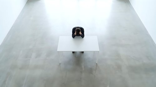 Lone businessman sitting at white desk in empty modern office space with polished concrete floors, minimalist workspace for corporate concepts.
