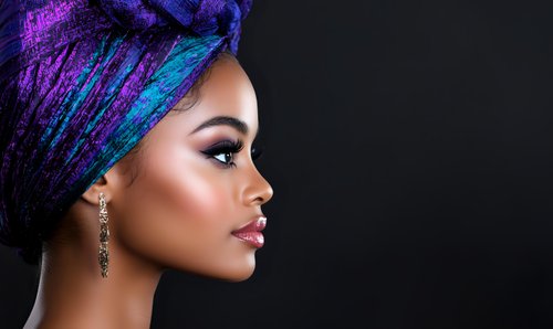 Young African woman in vibrant blue and purple head wrap poses in profile against dark background, showcasing elegant earrings and glowing makeup.