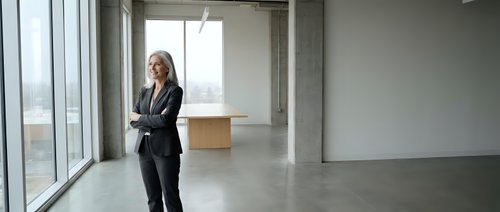 Businesswoman in formal black suit standing in empty modern office space with large windows and concrete flooring, suitable for corporate and professional business concepts.