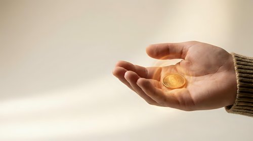 Hand holding glowing golden coin with warm light effect against soft beige background representing wealth, success, and financial opportunity.