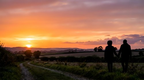 Two people standing on a rural path watching golden sunset over countryside landscape with mountains in distance, suitable for lifestyle and nature editorial use.