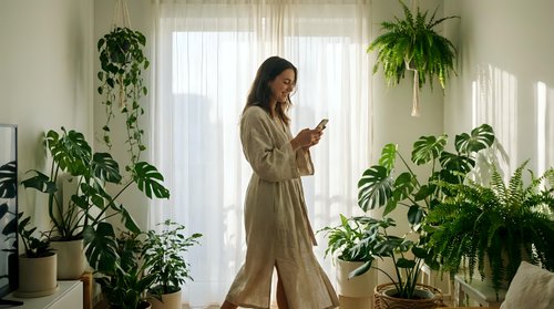 Young woman in linen outfit using smartphone among potted houseplants by bright window, surrounded by green foliage for home decor and lifestyle content.