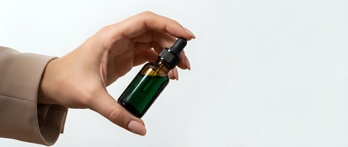 Hand holding small green glass dropper bottle with black cap against white background for skincare and cosmetic product presentation.