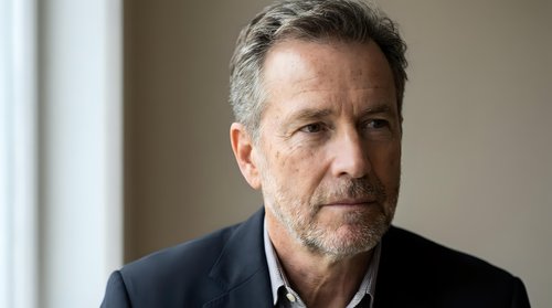 Middle-aged man with salt-and-pepper hair and facial stubble wearing dark blazer over white shirt. Professional portrait suitable for corporate, business, and editorial use.