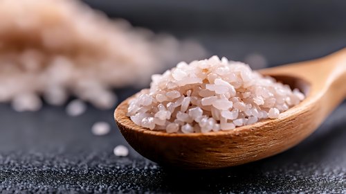 Pink Himalayan salt crystals in wooden spoon on dark background, macro shot with selective focus showing mineral texture and natural gourmet seasoning.