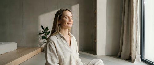 Young Caucasian woman in white bathrobe relaxing by window with natural sunlight in modern spa interior for wellness and self-care lifestyle.