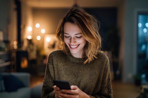 Young Caucasian woman with wavy hair smiling at smartphone in cozy evening home atmosphere with warm lighting and bokeh background.