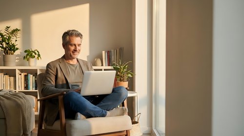 Mature Caucasian man working on laptop computer at home in bright living room with natural sunlight streaming through window creating warm atmosphere.