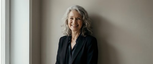 Confident mature Caucasian businesswoman with gray hair smiling in modern office interior wearing professional black blazer for corporate lifestyle.