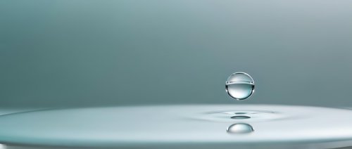 Single water droplet suspended above calm surface creating perfect sphere with reflection below against soft teal gradient background for wellness concepts.