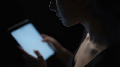 Young woman using tablet computer in dark room with glowing screen light illuminating her face for late night browsing and digital technology concepts.